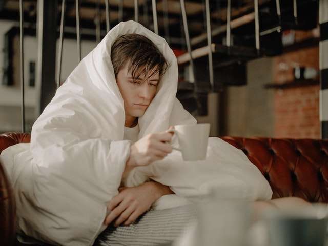 A man feeling sick under a blanket and holding a mug